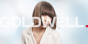 Woman with dark wavy hair wearing a white blouse in front of bright background and Goldwell logo in front